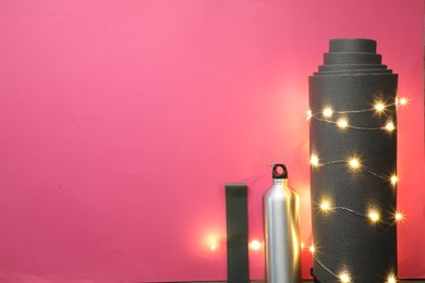 Photo of Yoga mat, band, bottle and Christmas lights on pink background, flat lay. Space for text