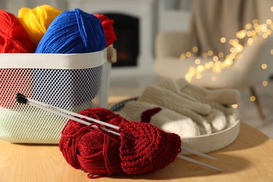 Colorful yarns, knitting needles, mittens and basket on wooden table indoors, closeup