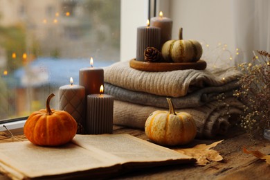 Photo of Burning candles, open book and stack of soft sweaters on windowsill. Autumn atmosphere