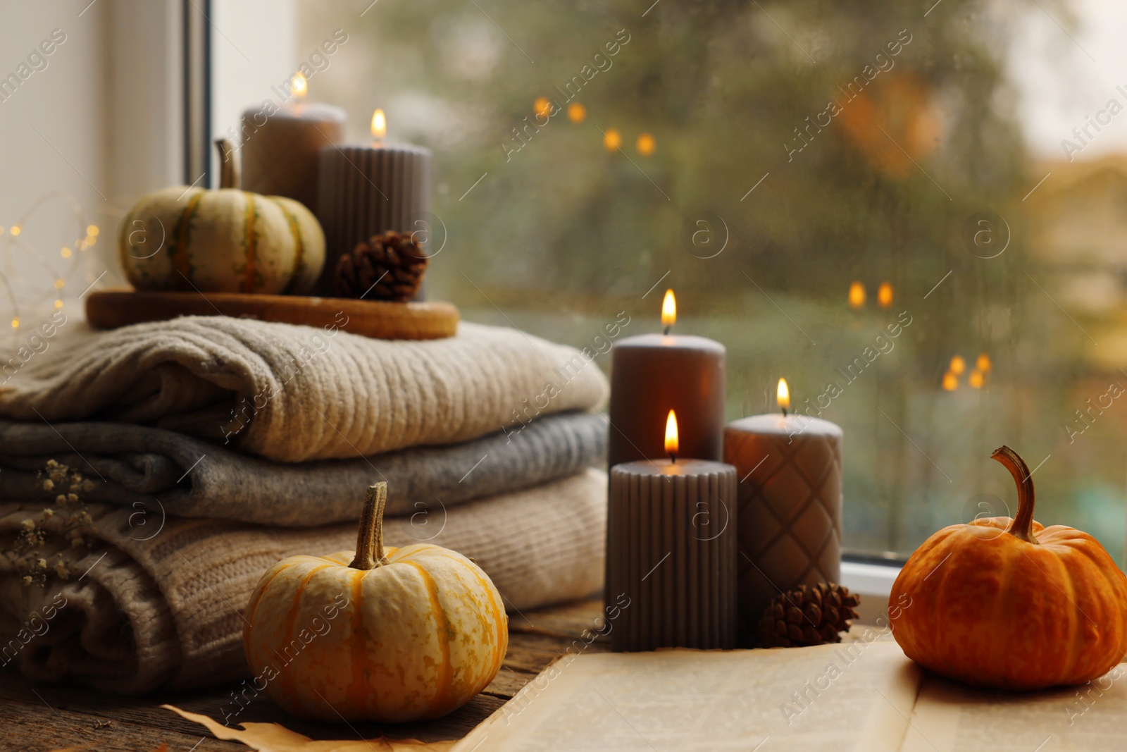 Photo of Burning candles, open book and stack of soft sweaters on windowsill. Autumn atmosphere