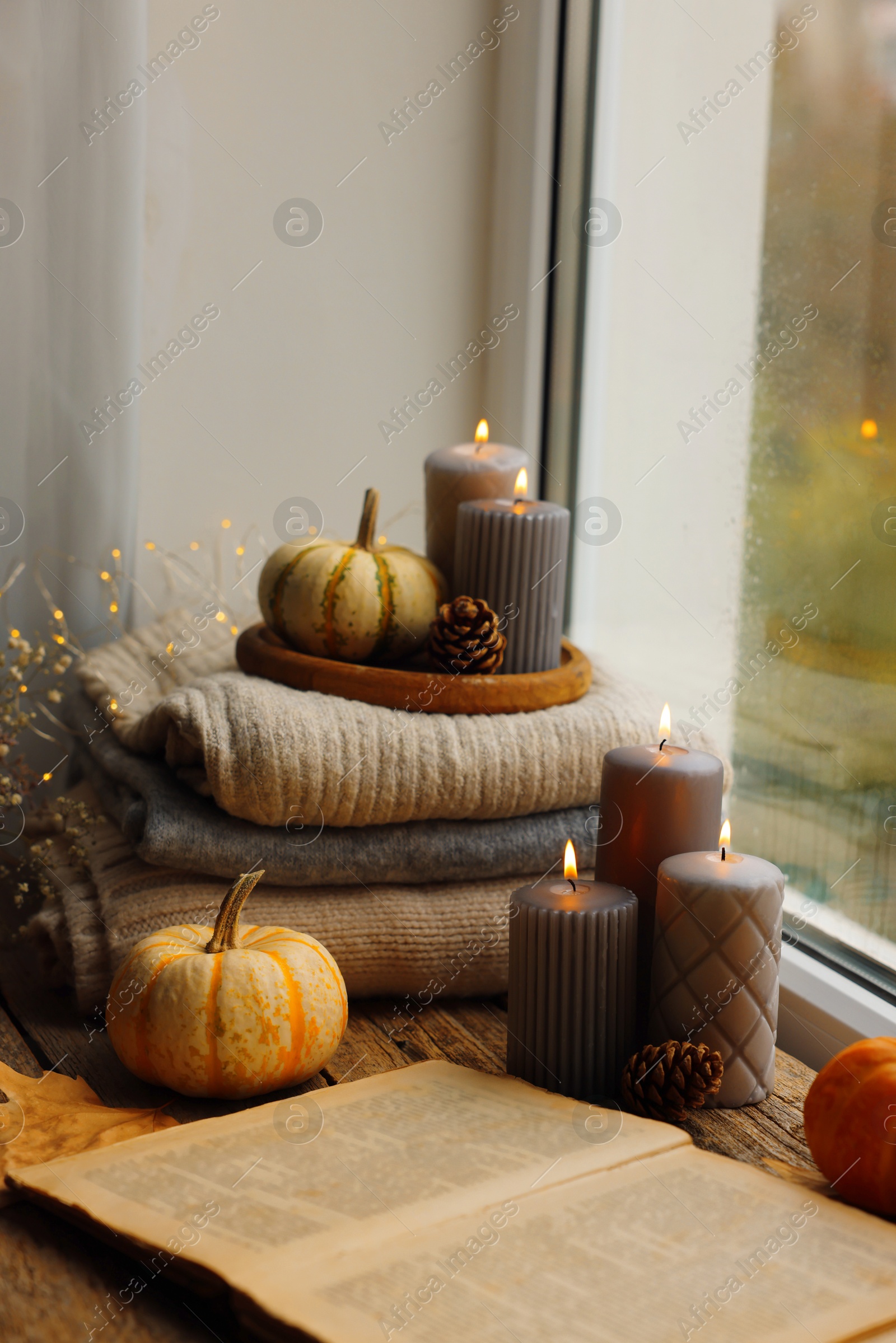 Photo of Burning candles, open book and stack of soft sweaters on windowsill. Autumn atmosphere