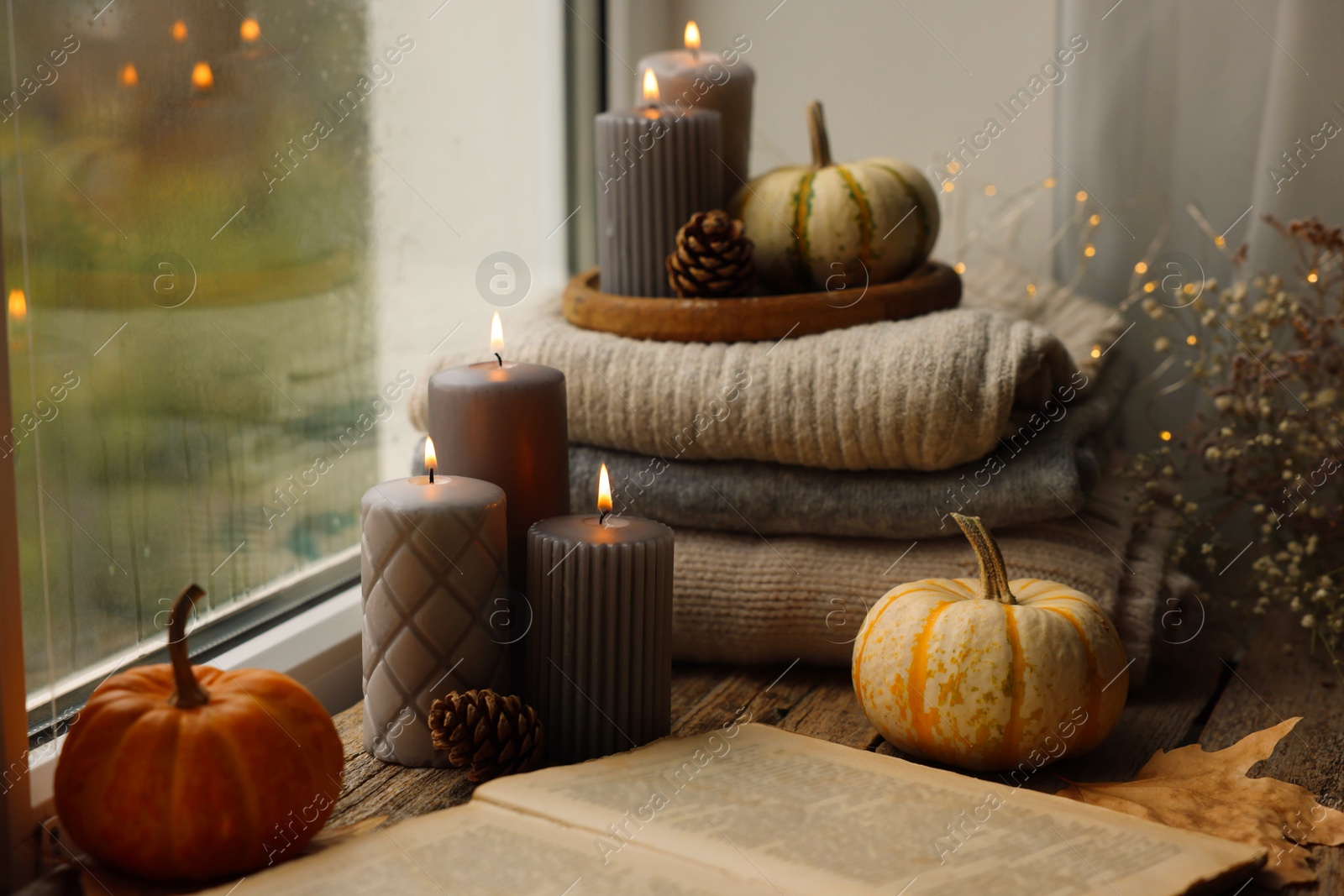 Photo of Burning candles, open book and stack of soft sweaters on windowsill. Autumn atmosphere