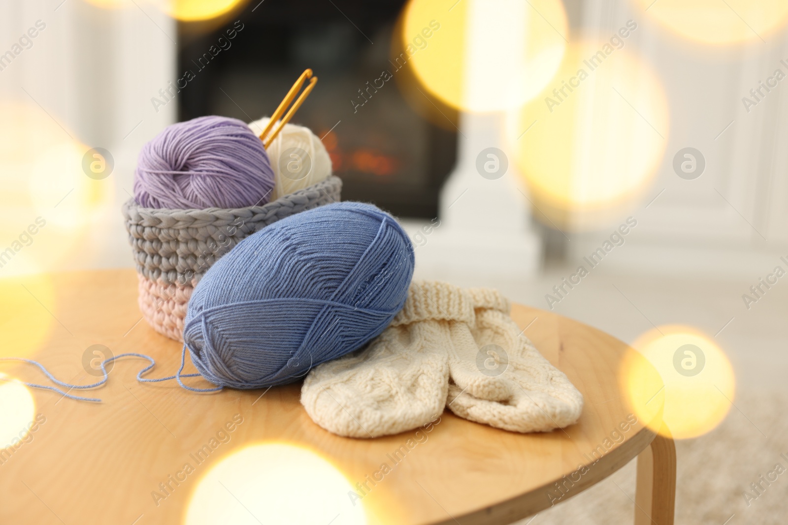 Photo of Colorful yarns, knitted mittens and crochet hooks on wooden table among blurred lights, closeup