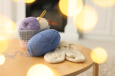 Photo of Colorful yarns, knitted mittens and crochet hooks on wooden table among blurred lights, closeup