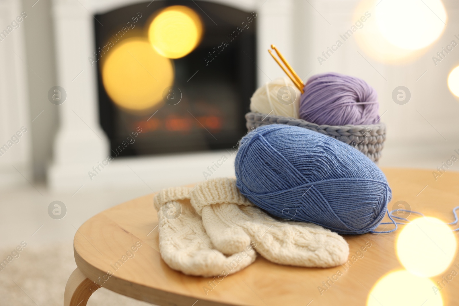 Photo of Colorful yarns, knitted mittens and crochet hooks on wooden table among blurred lights, closeup