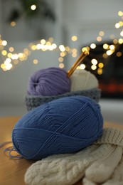 Photo of Colorful yarns, knitted mittens and crochet hooks on wooden table against blurred lights, closeup