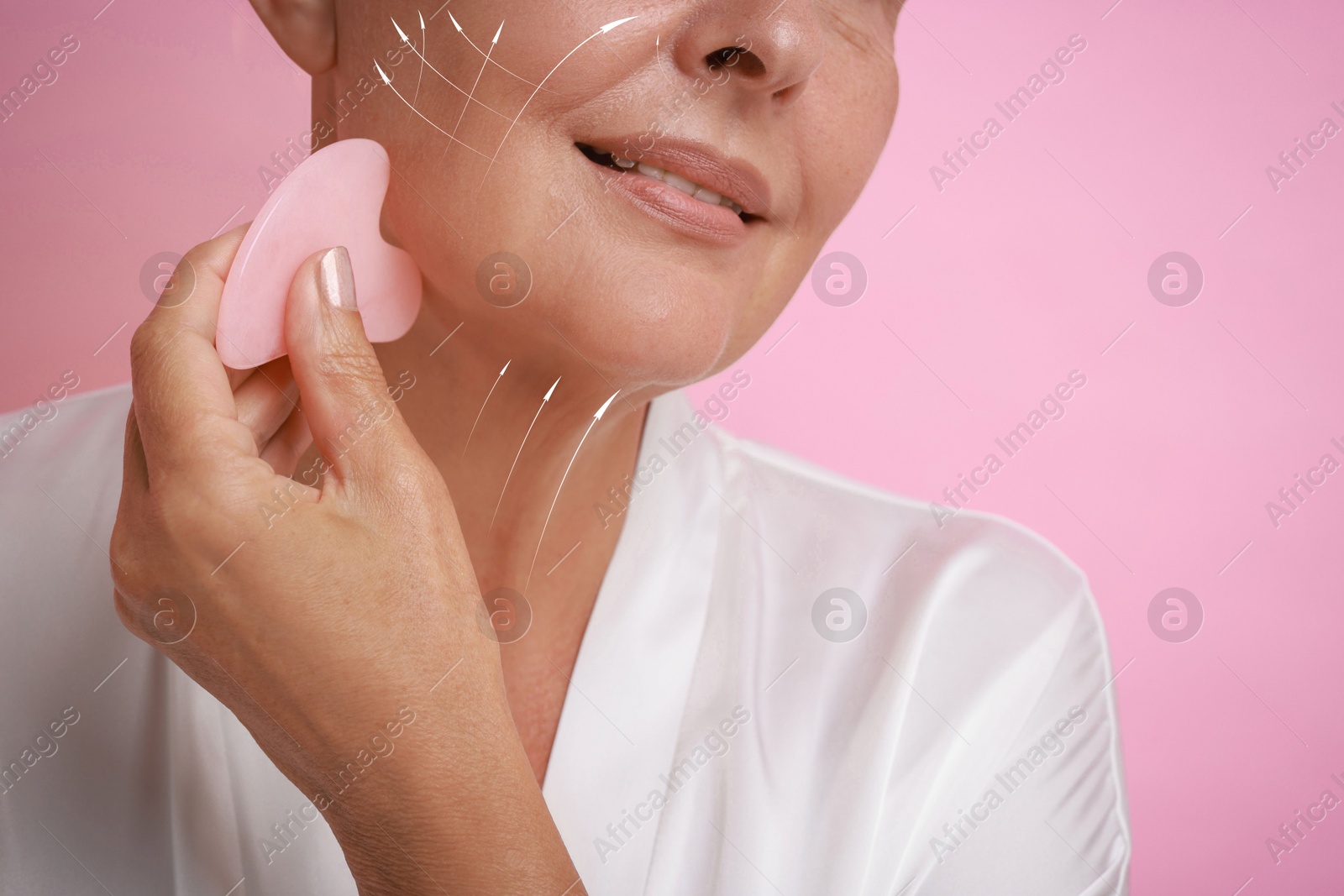 Image of Woman doing facial massage with gua sha tool on pink background, closeup. Arrows on skin showing direction of moves