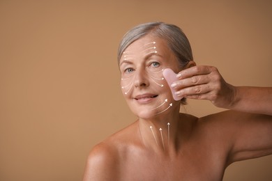 Beautiful woman doing facial massage with gua sha tool on pale brown background. Arrows on skin showing direction of moves