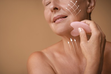 Image of Woman doing facial massage with gua sha tool on pale brown background, closeup. Arrows on skin showing direction of moves