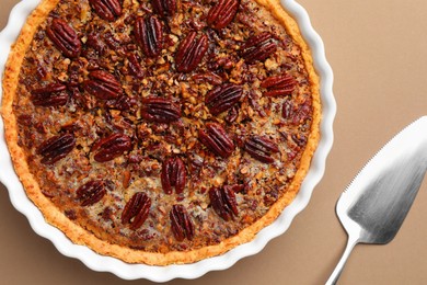 Photo of Delicious pecan pie in baking dish and cake server on light brown background, flat lay