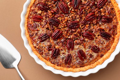 Photo of Delicious pecan pie in baking dish and cake server on light brown background, flat lay