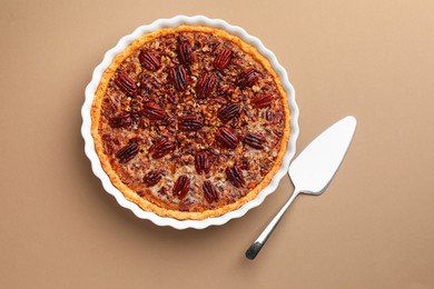 Photo of Delicious pecan pie in baking dish and cake server on light brown background, flat lay