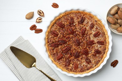 Photo of Delicious pecan pie in baking dish, fresh nuts and cake server on white wooden table, flat lay