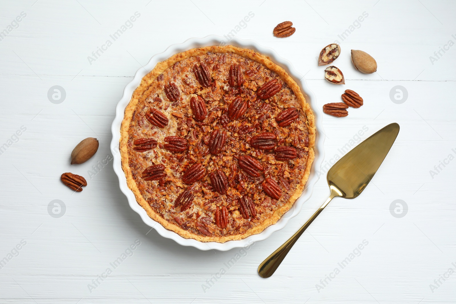 Photo of Delicious pecan pie in baking dish, cake server and fresh nuts on white wooden table, flat lay