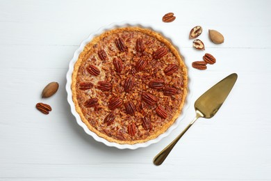 Delicious pecan pie in baking dish, cake server and fresh nuts on white wooden table, flat lay