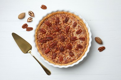 Photo of Delicious pecan pie in baking dish, cake server and fresh nuts on white wooden table, flat lay