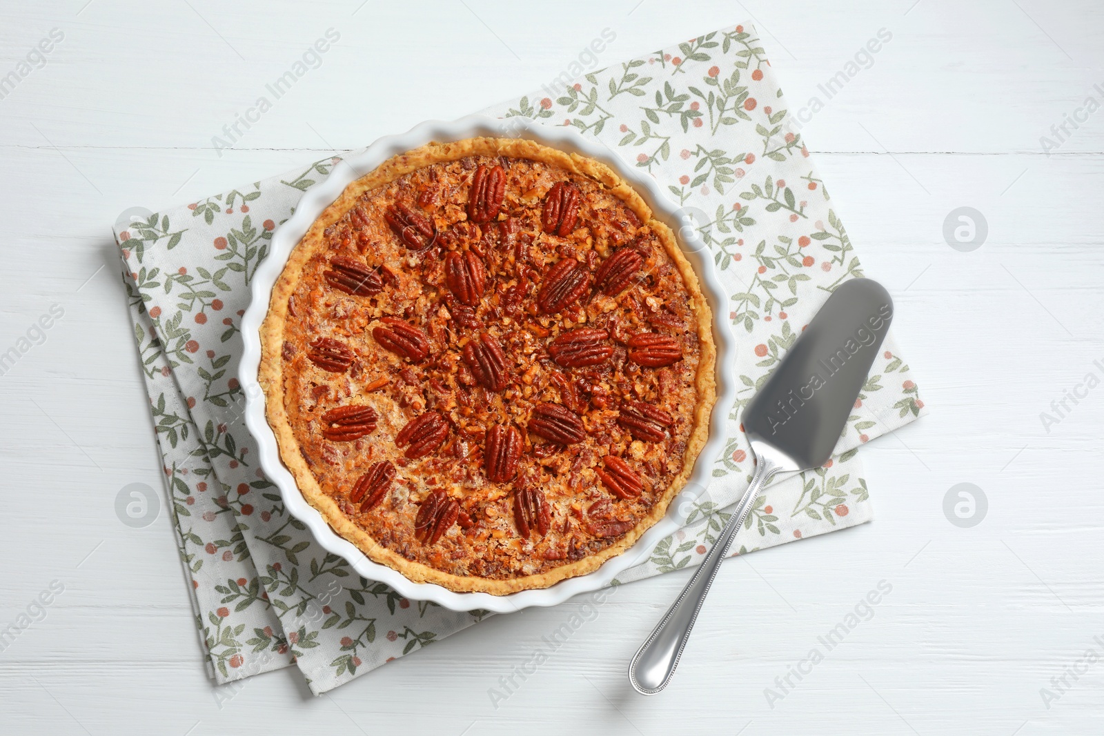 Photo of Delicious pecan pie in baking dish and cake server on white wooden table, flat lay