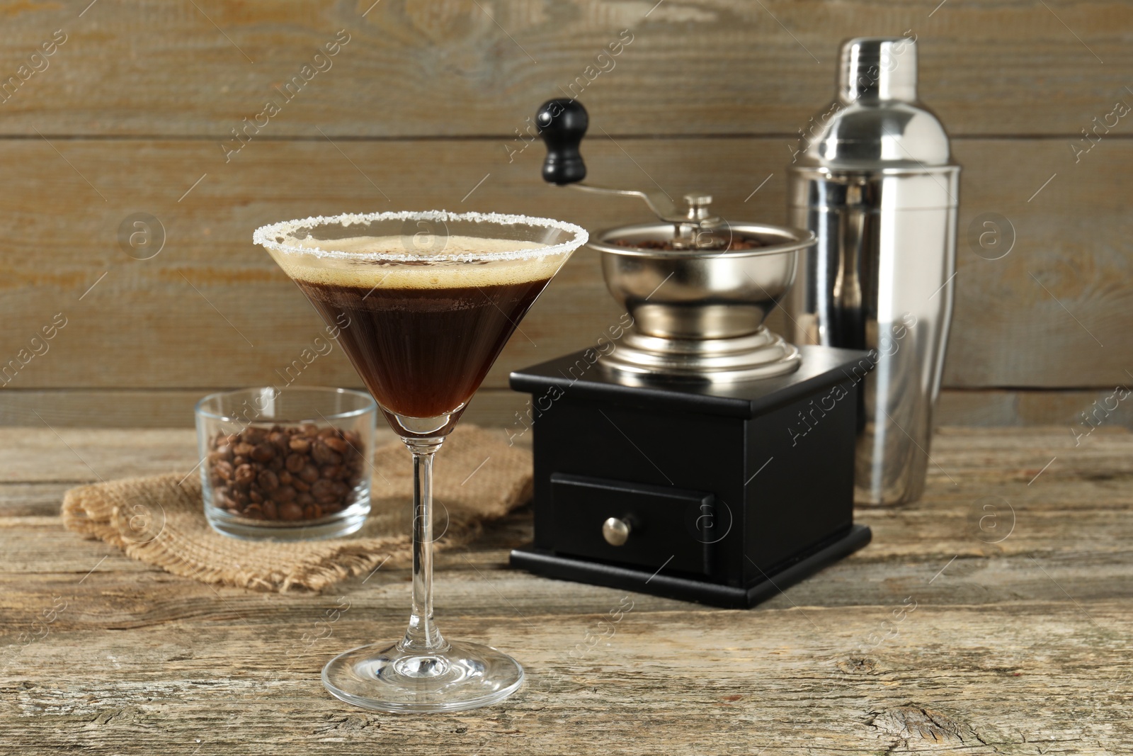 Photo of Glass of fresh cocktail, manual coffee grinder, shaker and beans in bowl on wooden table