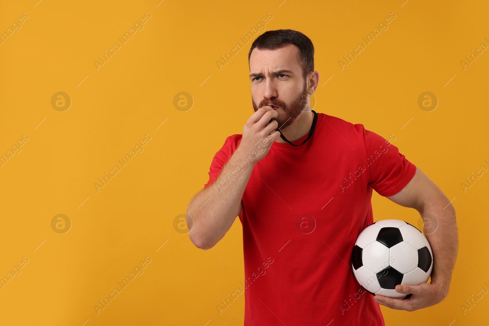 Photo of Young man with soccer ball blowing whistle on orange background, space for text