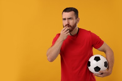 Young man with soccer ball blowing whistle on orange background, space for text