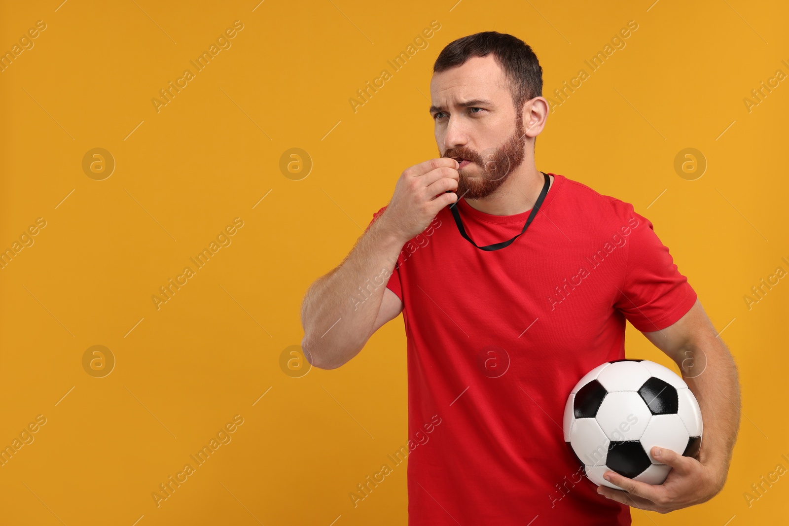 Photo of Young man with soccer ball blowing whistle on orange background, space for text