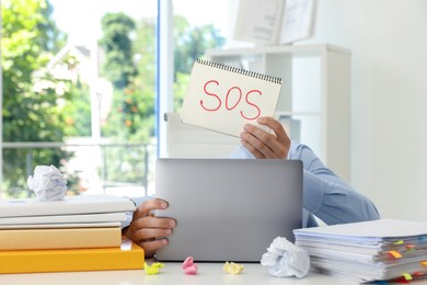 Photo of Man holding notebook with word SOS and hiding face behind laptop at table in office