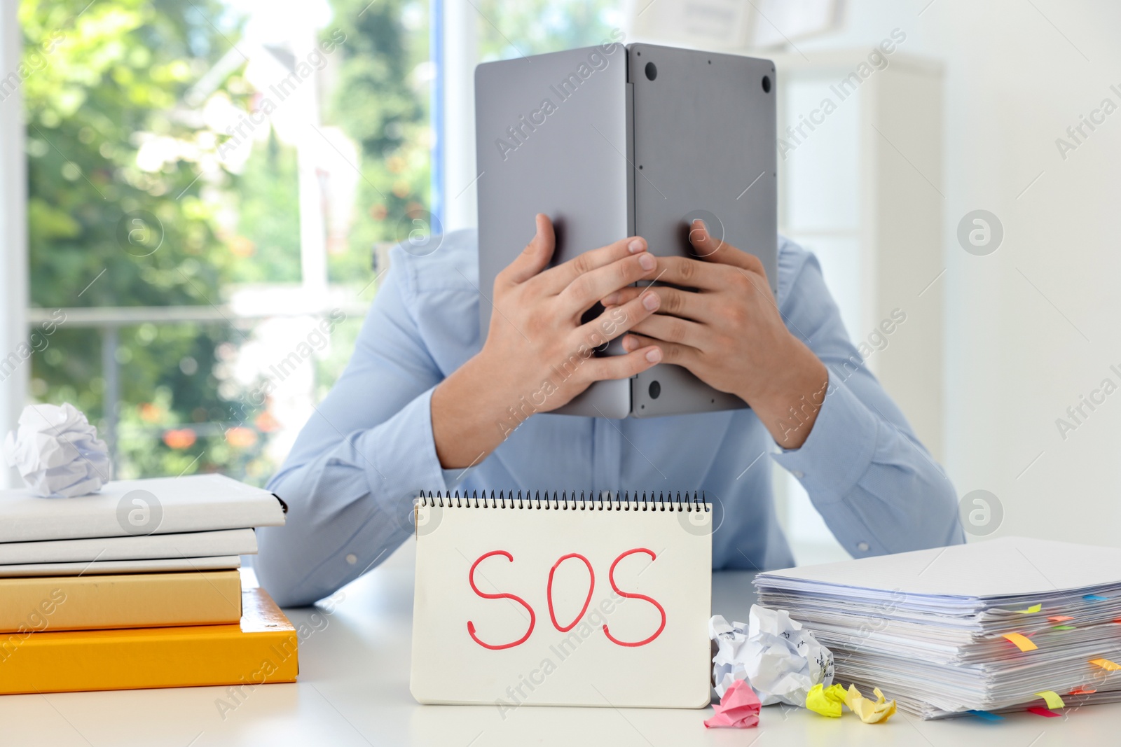 Photo of Man hiding face behind laptop near notebook with word SOS at table in office