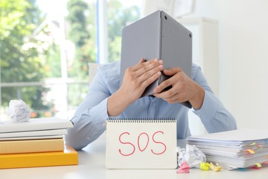 Photo of Man hiding face behind laptop near notebook with word SOS at table in office