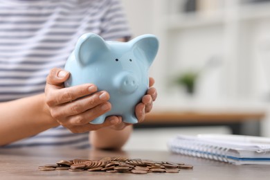 Photo of Woman holding piggy bank over table with coins indoors, closeup