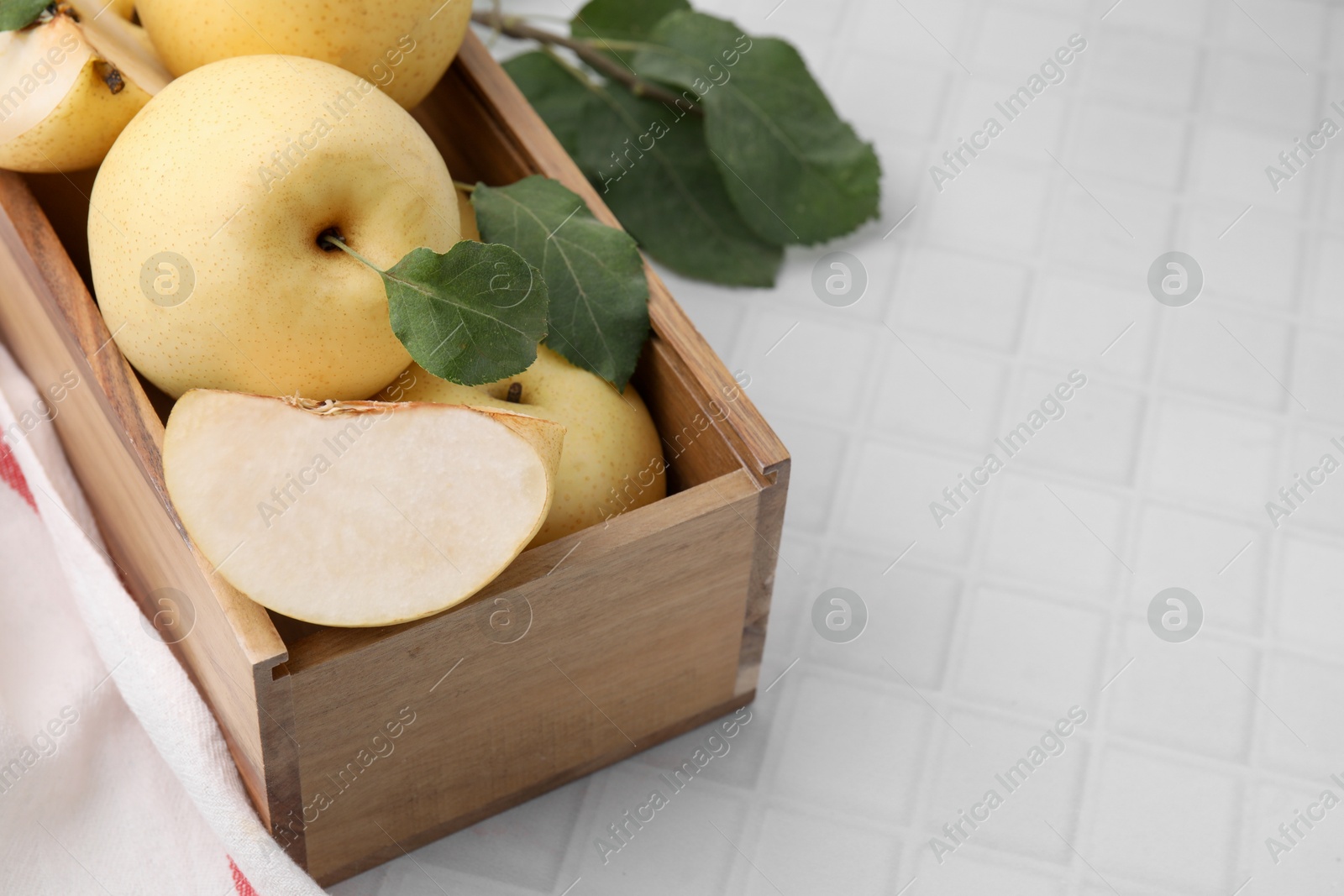 Photo of Delicious fresh apple pears in wooden crate and green leaves on white tiled table, space for text