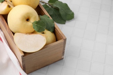 Photo of Delicious fresh apple pears in wooden crate and green leaves on white tiled table, space for text