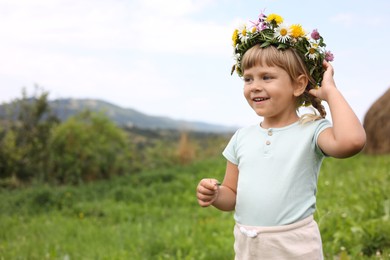 Photo of Portrait of smiling little girl in floral wreath at green meadow, space for text. Child enjoying beautiful nature