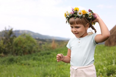 Portrait of cute little girl in floral wreath at green meadow, space for text. Child enjoying beautiful nature