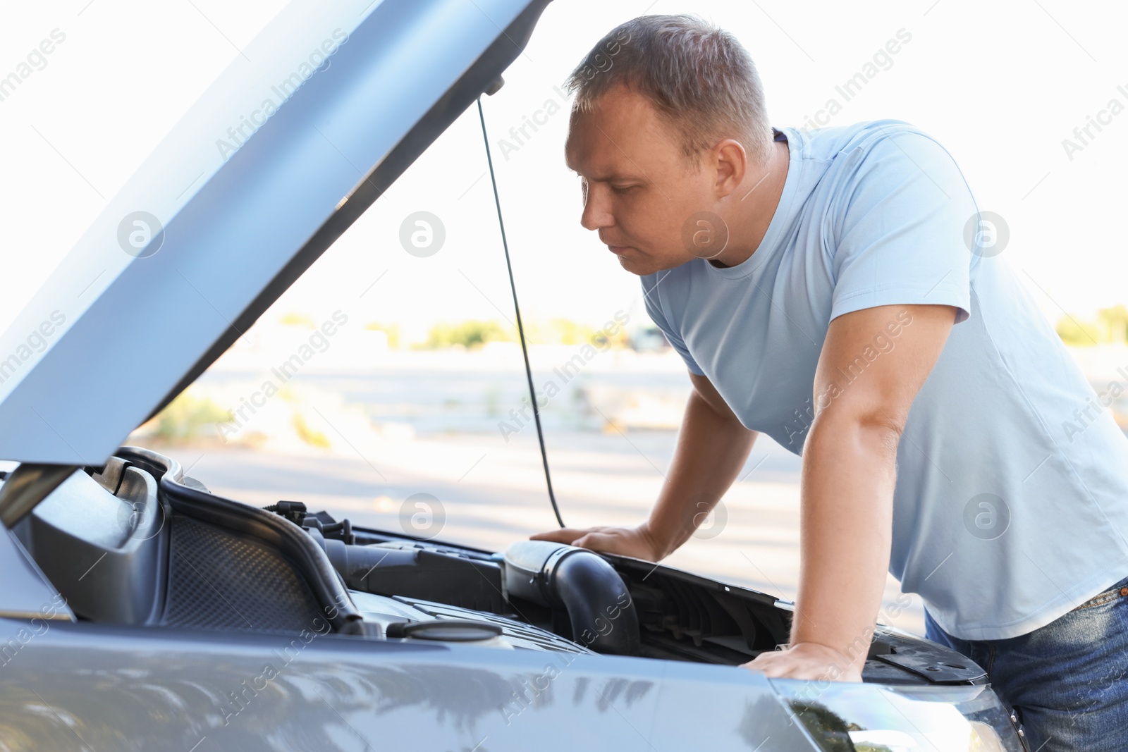 Photo of Stressed man looking under hood of broken car outdoors