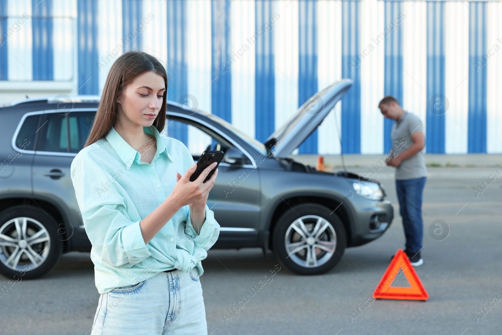 Photo of Man standing near broken car while woman using smartphone outdoors