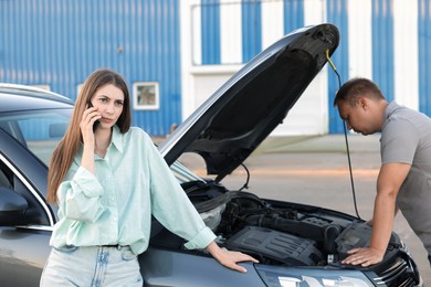 Man examining broken auto while woman calling to car service outdoors