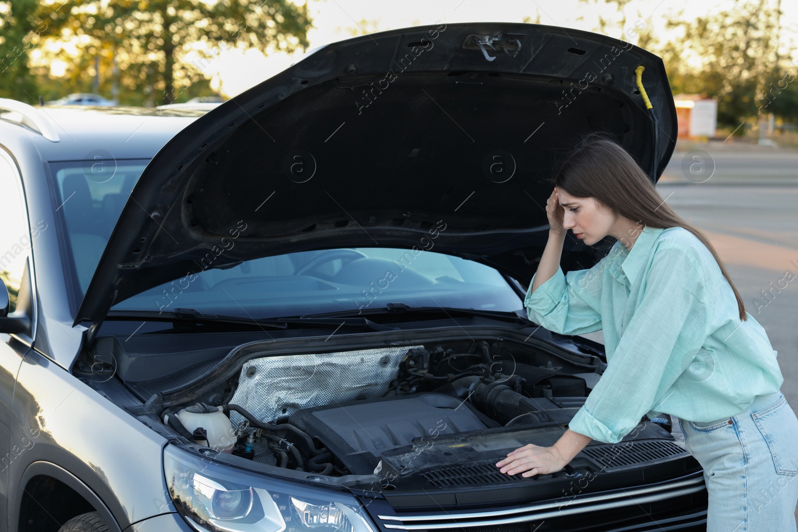 Photo of Stressed woman looking under hood of broken car outdoors