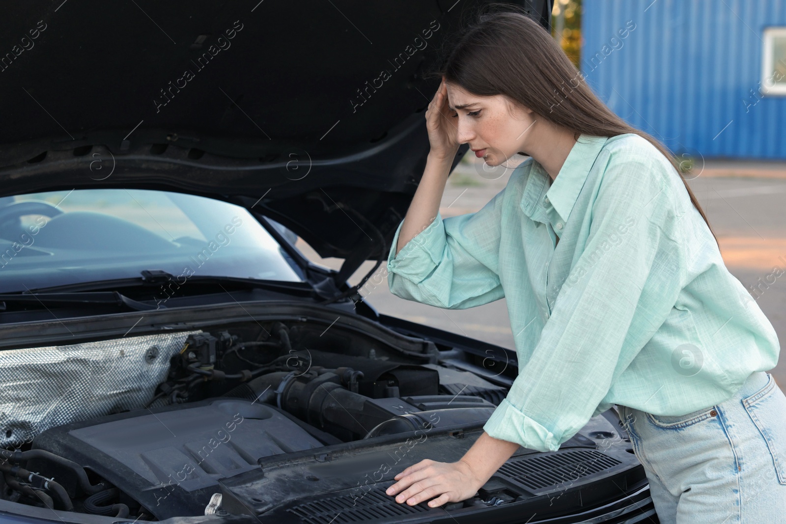 Photo of Stressed woman looking under hood of broken car outdoors