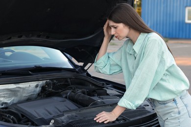 Photo of Stressed woman looking under hood of broken car outdoors