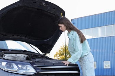 Photo of Stressed woman looking under hood of broken car outdoors