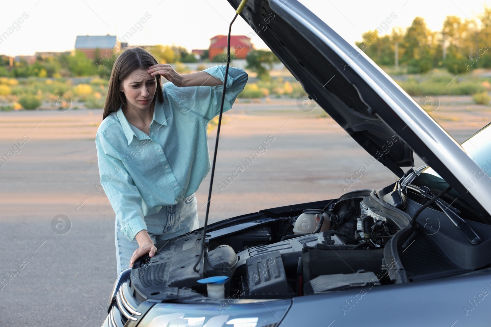 Photo of Stressed woman looking under hood of broken car outdoors