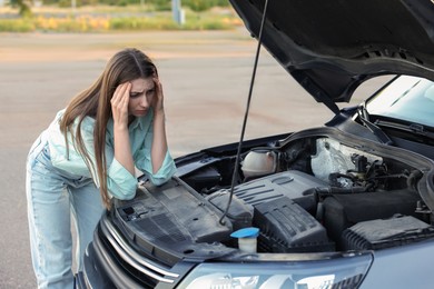 Photo of Stressed woman looking under hood of broken car outdoors