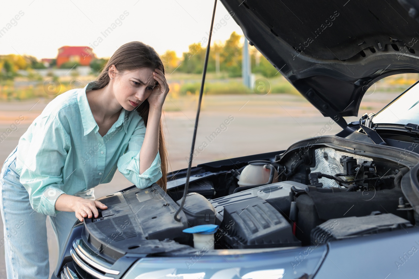 Photo of Stressed woman looking under hood of broken car outdoors