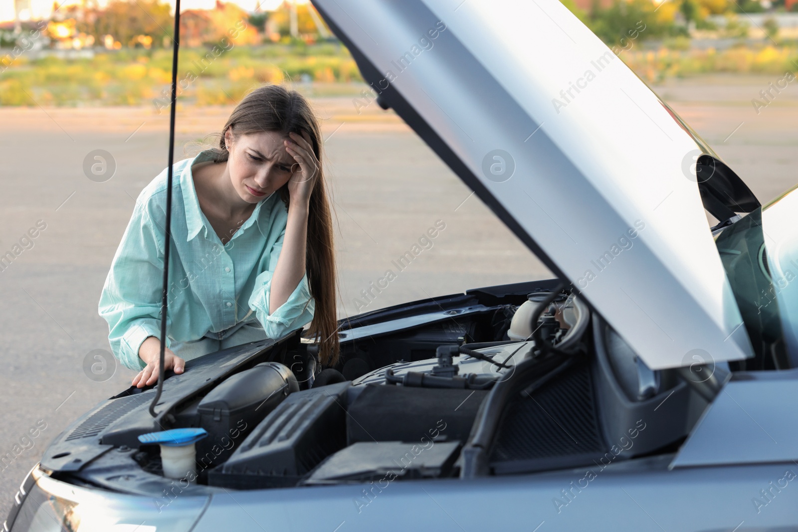 Photo of Stressed woman looking under hood of broken car outdoors