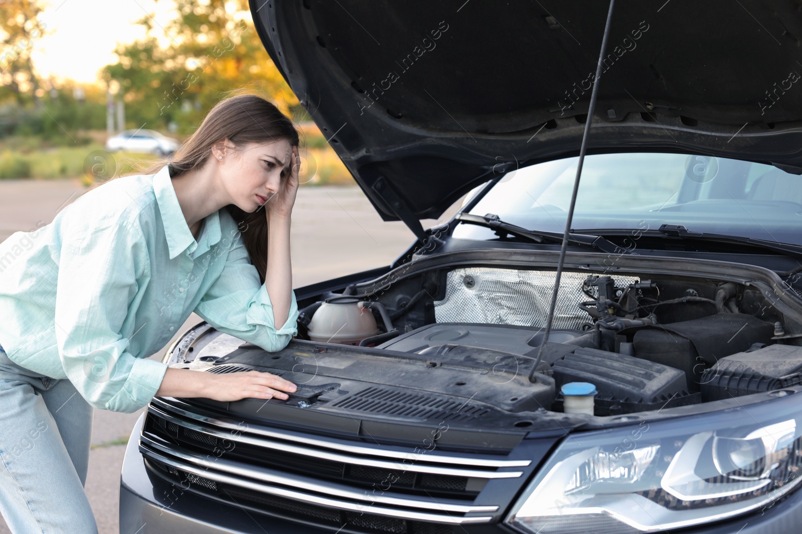 Photo of Stressed woman looking under hood of broken car outdoors