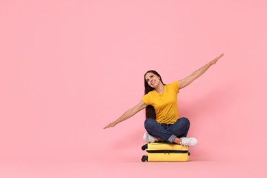 Happy young woman sitting on suitcase against pink background, space for text