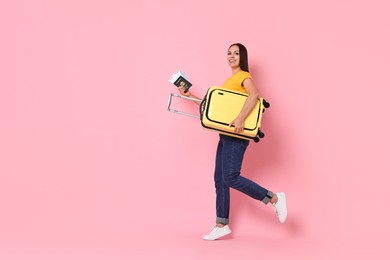 Happy young woman with suitcase, passport and ticket walking on pink background