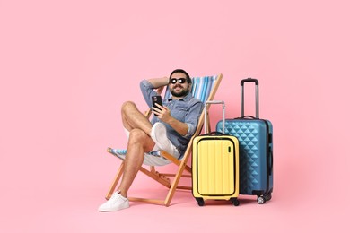 Happy man in sunglasses with smartphone sitting on folding chair and suitcases against pink background