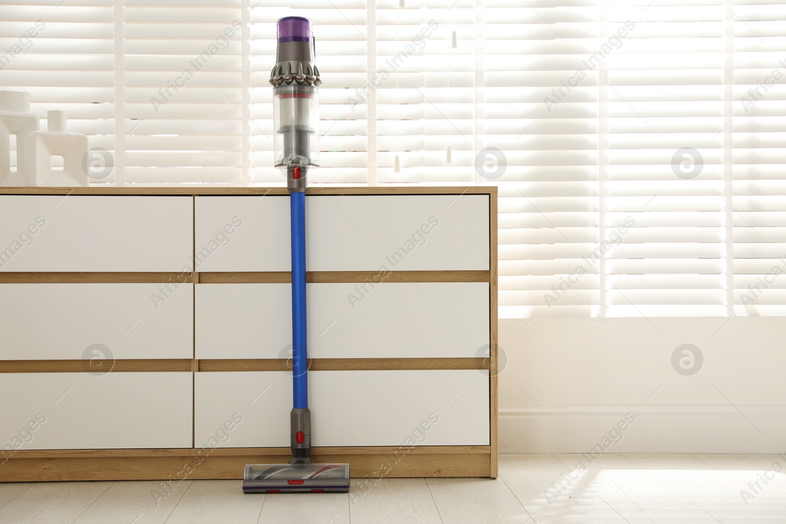 Photo of One cordless vacuum cleaner leaning on chest of drawers indoors