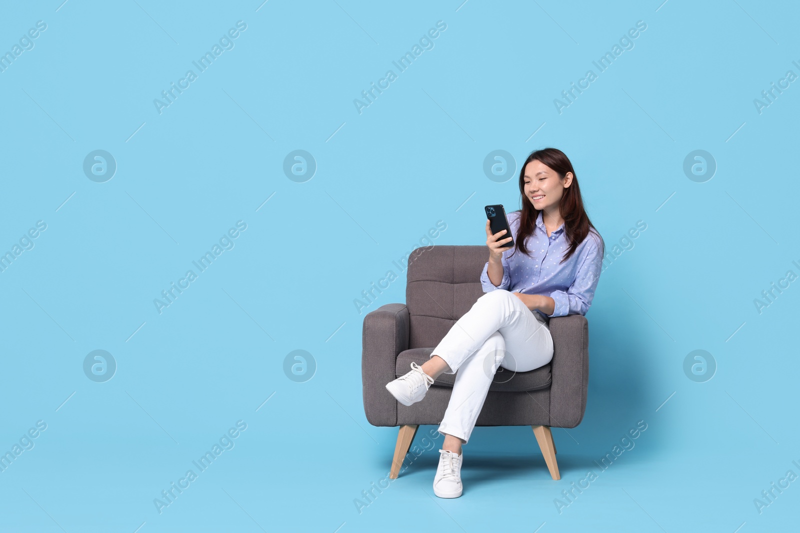 Photo of Smiling woman with smartphone resting in armchair on light blue background. Space for text